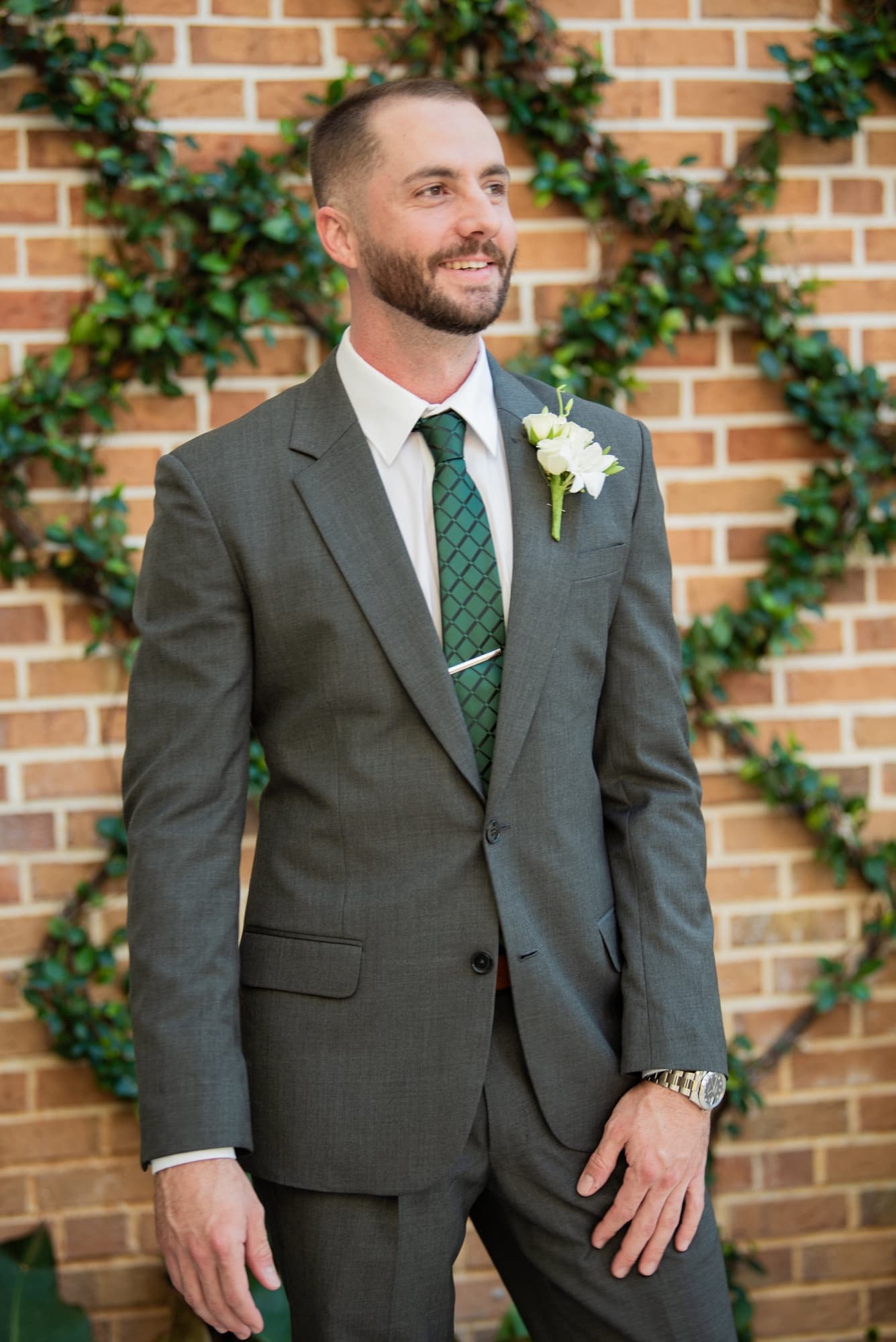 Groom in a grey-green suit with boutonnière against brick and ivy